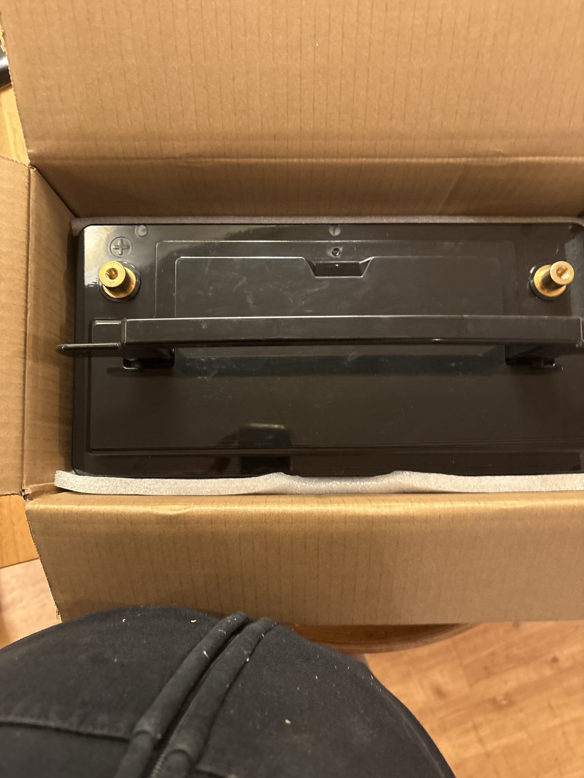 Schwarze Ultimatron-LiFePO4-Batterie von oben im geöffneten braunen Versandkarton. Die beiden messingfarbenen Batterieanschlüsse sind an den oberen Ecken sichtbar.