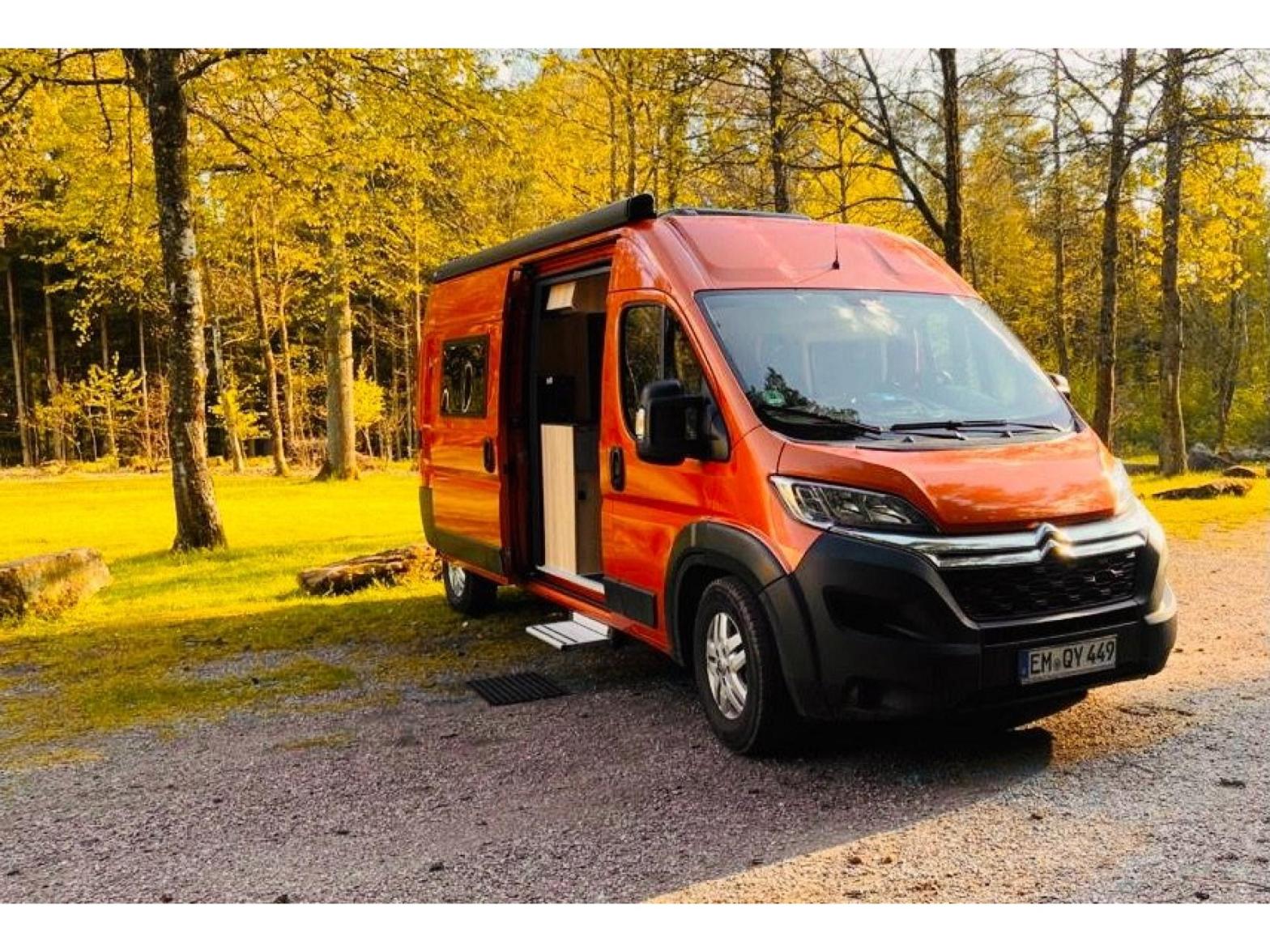 Der orangefarbene Kastenwagen Carl steht mit geöffneter Schiebetür und ausgefahrener Trittstufe auf einem Stellplatz vor goldgelben Laubbäumen.